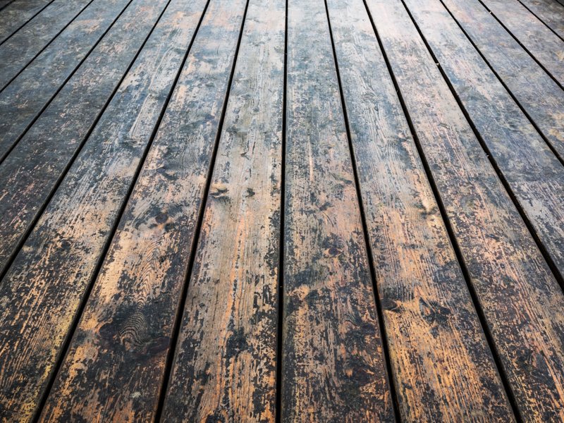 Close-up of a weathered wooden deck or floor made of long, narrow planks arranged in parallel. The wood shows signs of age, with visible scratches, stains, knots, and discoloration