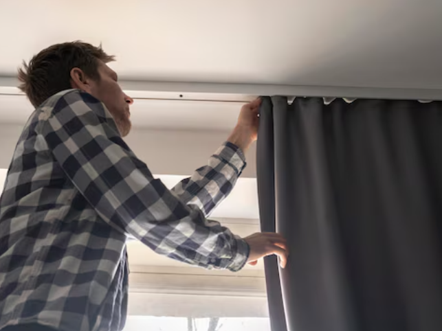 Person wearing a checkered shirt installing a dark curtain on a ceiling-mounted track near a window. They are reaching up to secure curtain hooks into the track, with natural light streaming through the window.