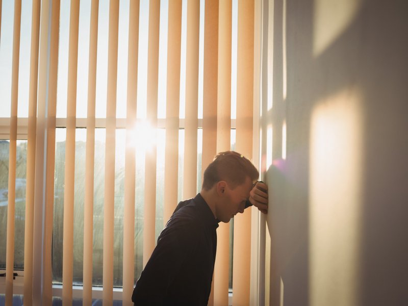 Person standing indoors beside a wall with vertical blinds covering large windows. They lean forward, resting their forehead and hand against the wall as sunlight filters through the blinds, casting warm shadows.